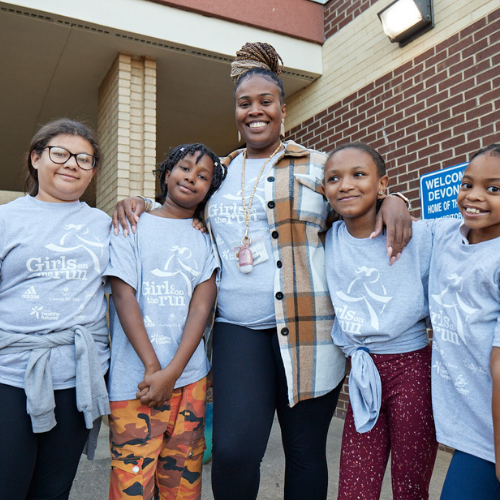 A bright outdoor photograph of four young people standing closely together and smiling at the camera. They are all wearing matching bright pink Girls on the Run t-shirts, and one person in the front has a white race bib attached to their shirt.